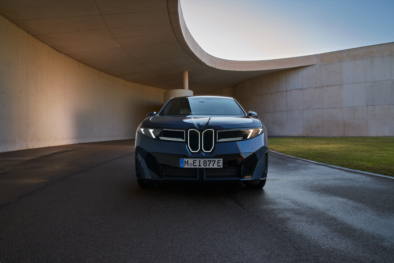 Ein blauer BMW iX3 steht frontal zur Kamera gerichtet auf einem Platz. Er ist von zwei Betonw&auml;nden und einem halb offenem Betondach umgeben.