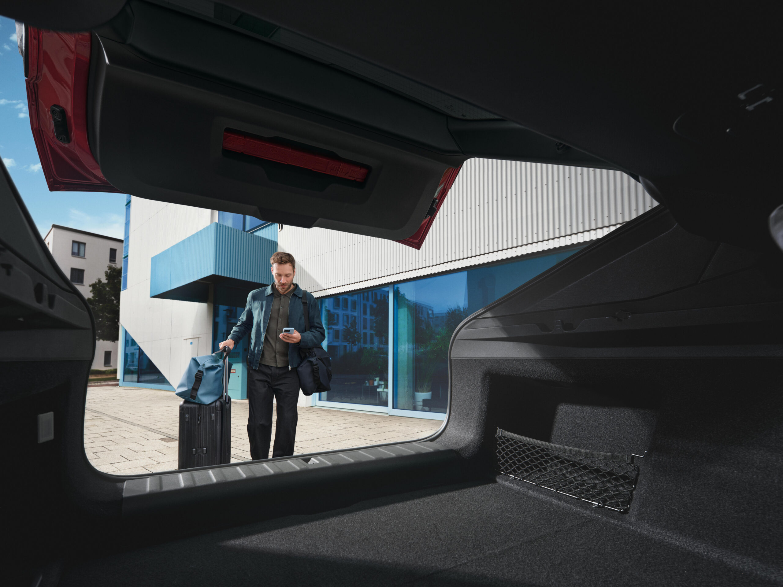 Mann mit KOffer und Handy in der Hand wird von einer Kamera in einem Autokofferraum heraus fotografiert, als er auf den ge&ouml;ffneten Kofferraum zugeht.