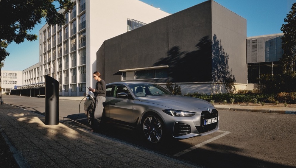 Ein silbener BMW i4 steht am Stra&szlig;enrand neben einer E-Lades&auml;ule. W&auml;hrend das Fahrzeug l&auml;d lehnt sich die Fahrerin des Wagens an das Auto und wartet.