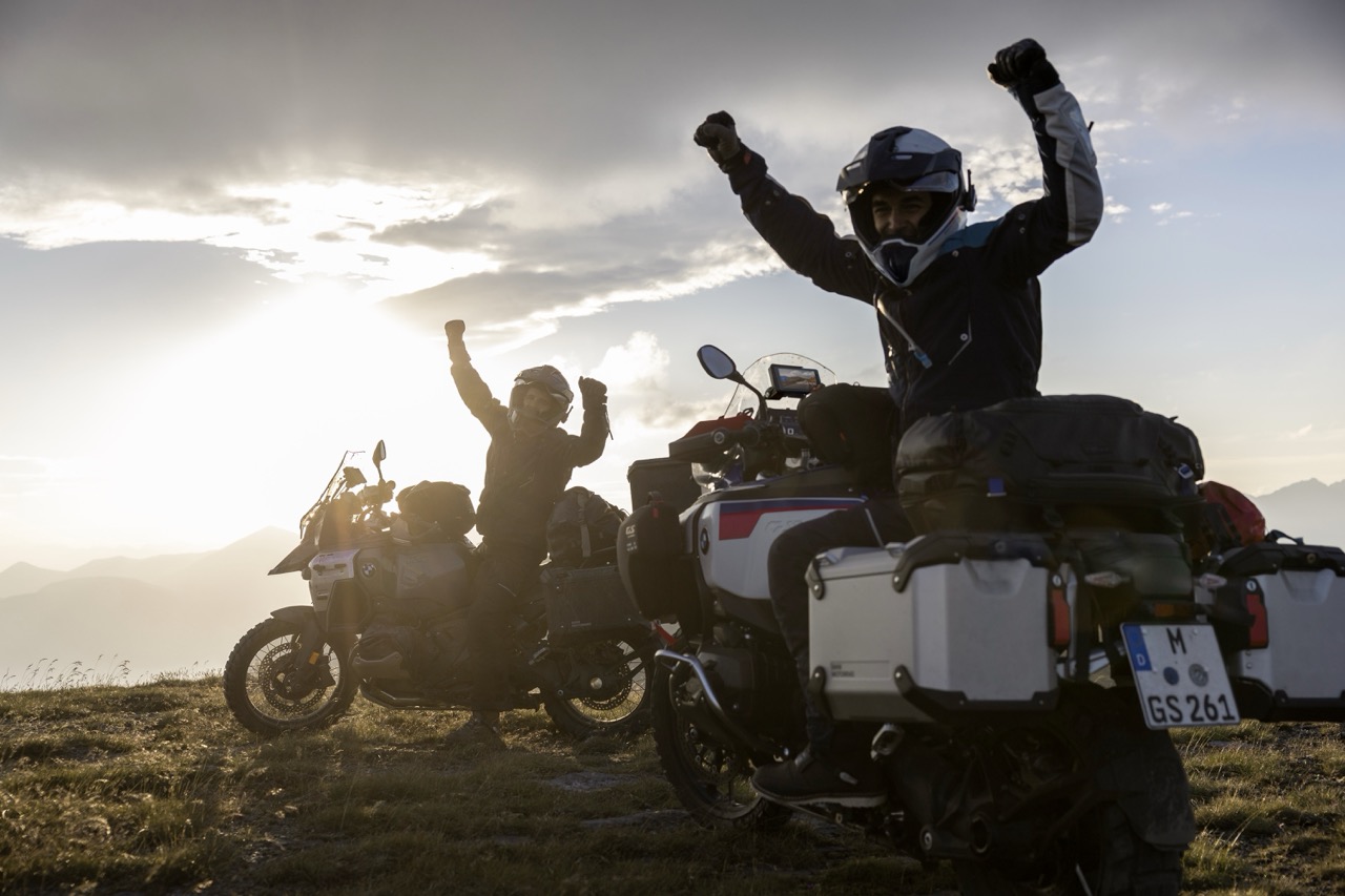 Zwei Motorradfahrer mit Helm und Fahrerausstattung auf ihren Bikes vor einem Sonnenuntergang. Sie drehen sich zur Kamera und jubeln.
