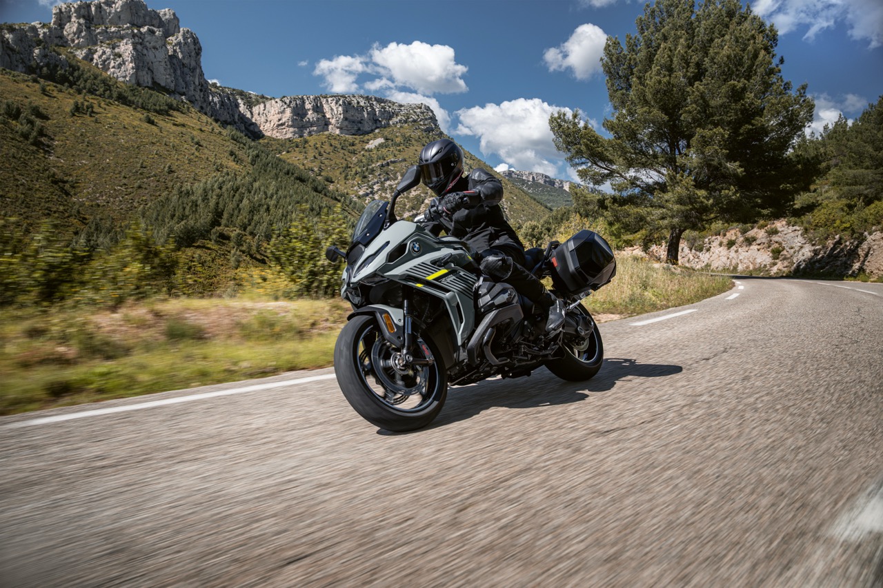 Motorradfahrer auf einer BMW R 1300 RS auf einem Bergpass unterwegs. Im Hintergrund sind grün bewachsene Berge zu sehen und ein strahlend blauer Himmel