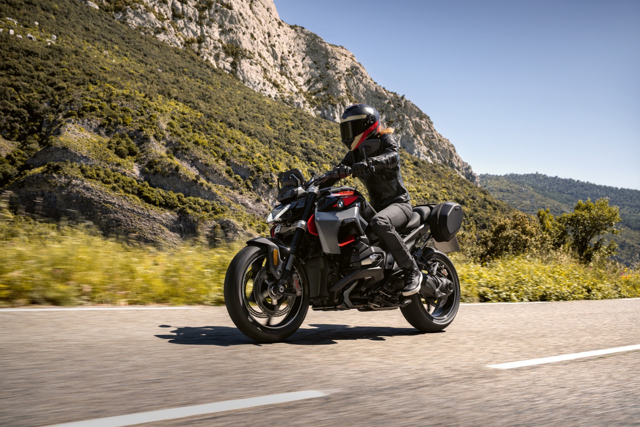 Motorradfahrer auf einer BMW R 1300 R auf einem Bergpass unterwegs. Im Hintergrund sind grün bewachsene Berge zu sehen und ein strahlend blauer Himmel