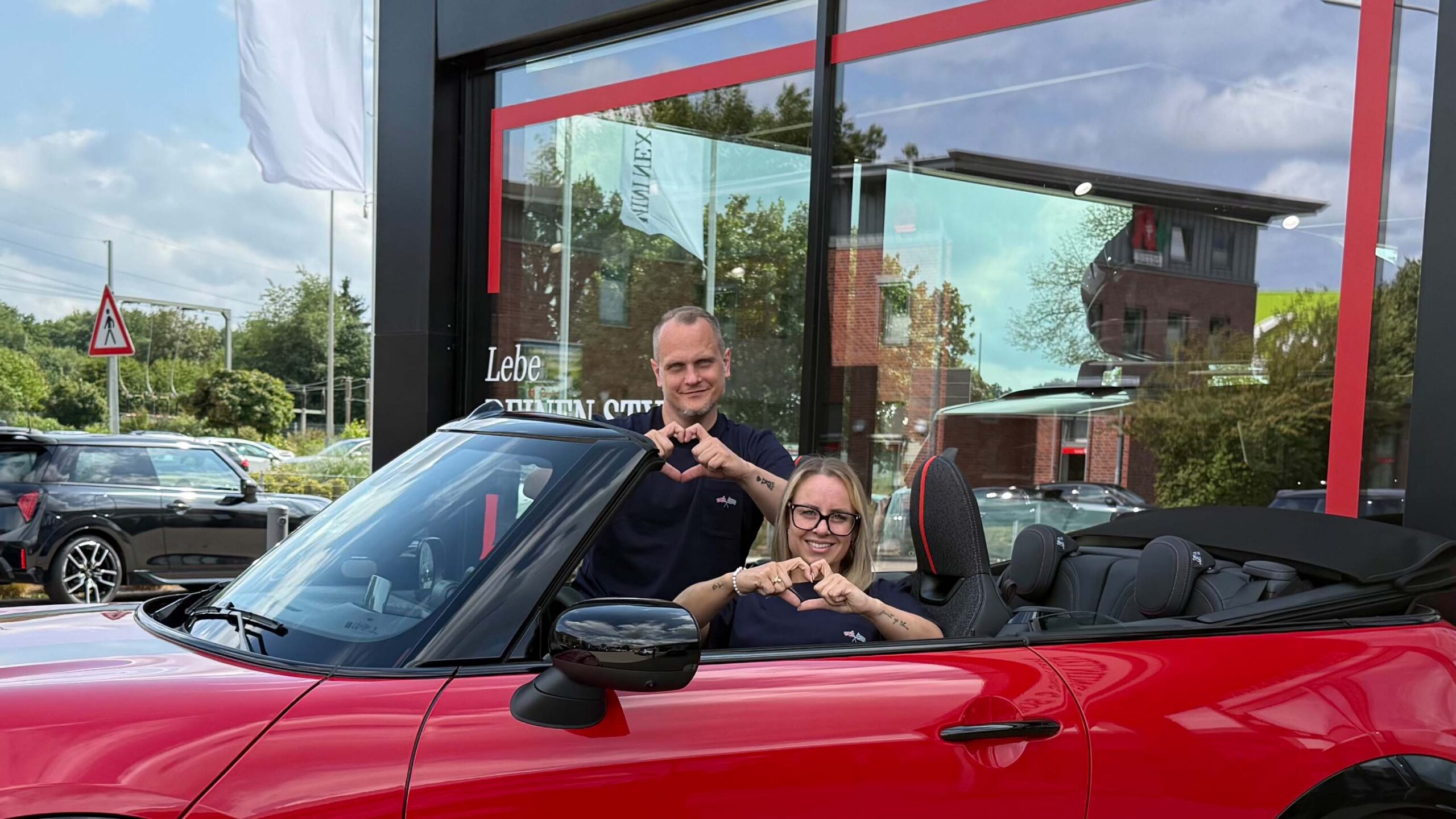Sarah und Jens sitzen im MINI Cabrio und zeigen Herzen in die Kamera