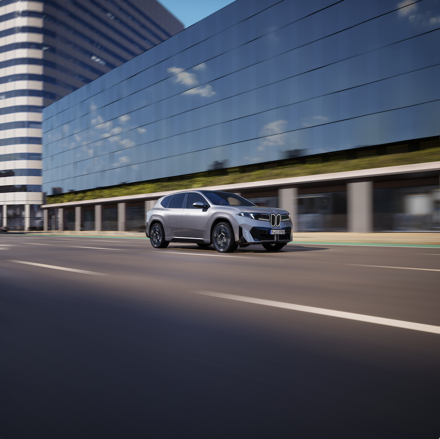 Zu sehen ist ein silberner BMW iX3, der auf einer Stra&szlig;e f&auml;hrt. Im Hintergrund sieht man ein gro&szlig;es Geb&auml;ude mit einer Glasfront.