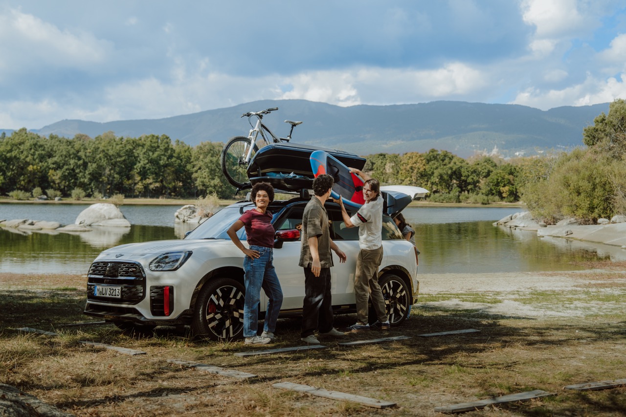 MINI Cooper steht bepackt mit Fahrrad, Dachbox und Surfbrett vor eine See. Davor stehen drei Menschen und entladen den MINI Cooper.