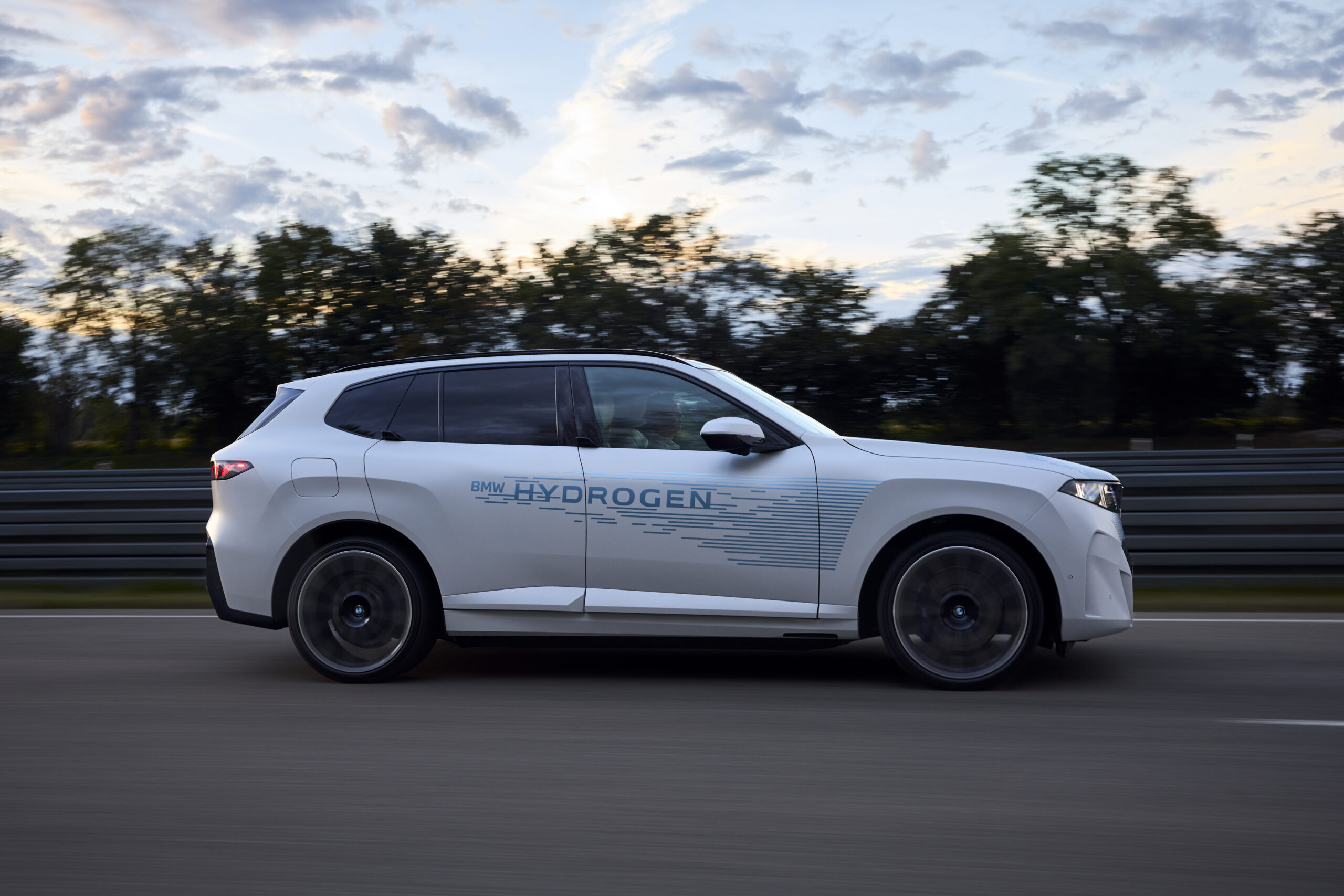 Man sieht einen weißen BMW iX5, der vollelektrisch und mit Wasserstoffantrieb auf der Straße fährt. Im Hintergrund sieht man einen Wald.
